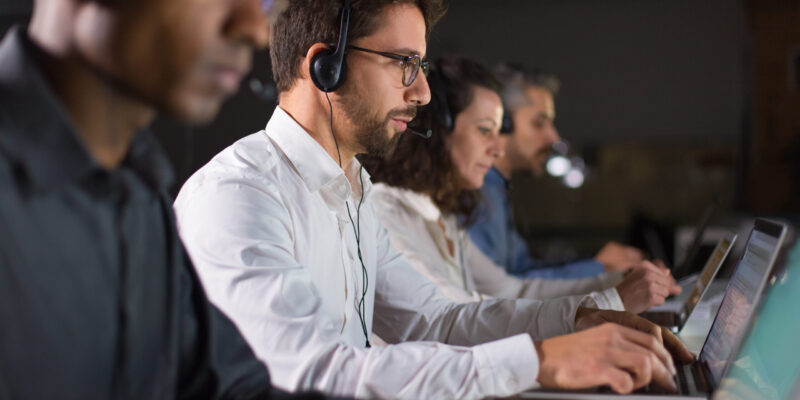 Side view of confident call center operator talking with client. Caucasian young man in eyeglasses typing on laptop while serving client. Call center concept