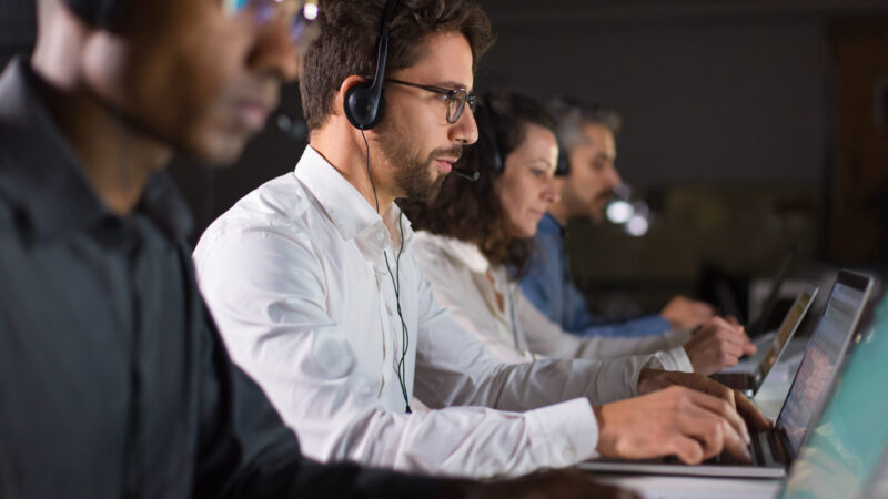 Side view of confident call center operator talking with client. Caucasian young man in eyeglasses typing on laptop while serving client. Call center concept